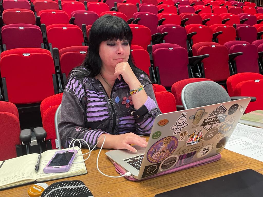 A white women with long dark haits sits in a theatre at a desk, at her laptop.