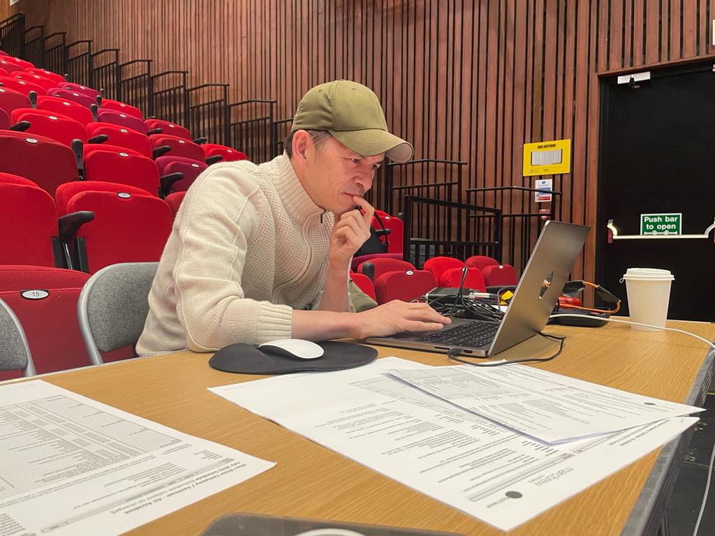 A white man wearing a baseball cap seated in a theatre at a desk leans over his laptop concentrating on the screen.
