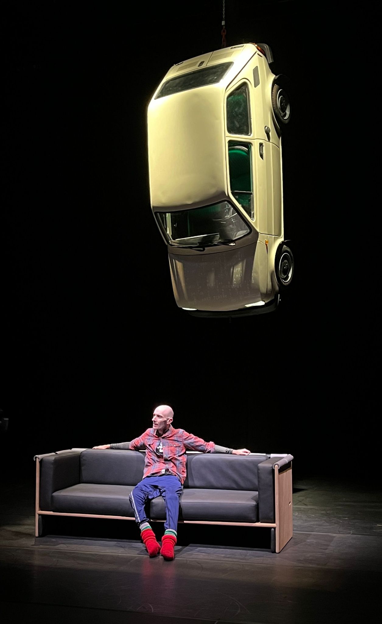 marc sits on a grey sofa, arms stretched along the back of it. The back drop is black. Above his head, suspended in the air is a small white car, illuminating him with its headlights.