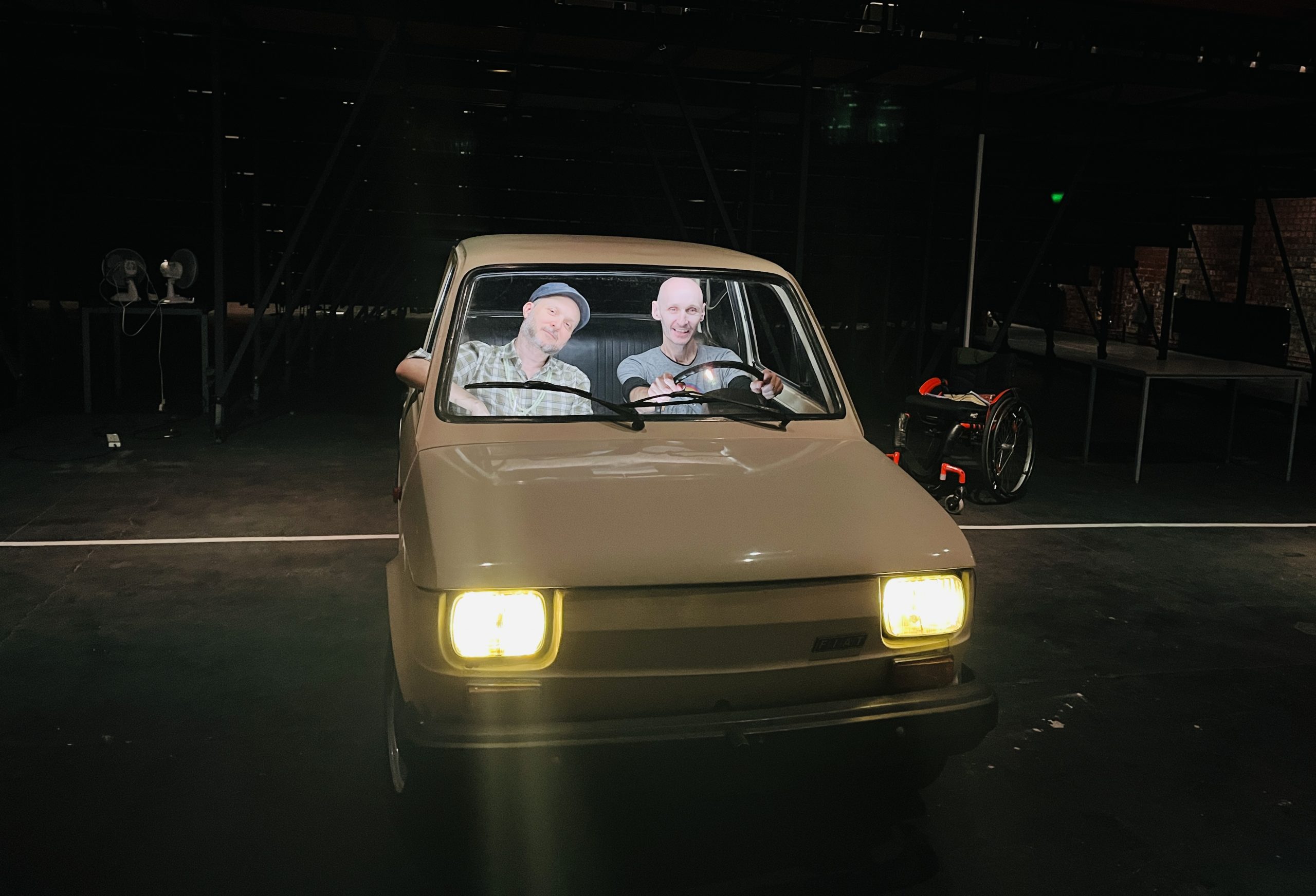 Sidi Larbi Cherkaoui sits in the passenger seat of a small white care. Marc is in the drivers seat. They're smiling. The cars headlights are lit. They're on a stage with a black dance floor. We can see Marc's wheelchair in the background behind them.  