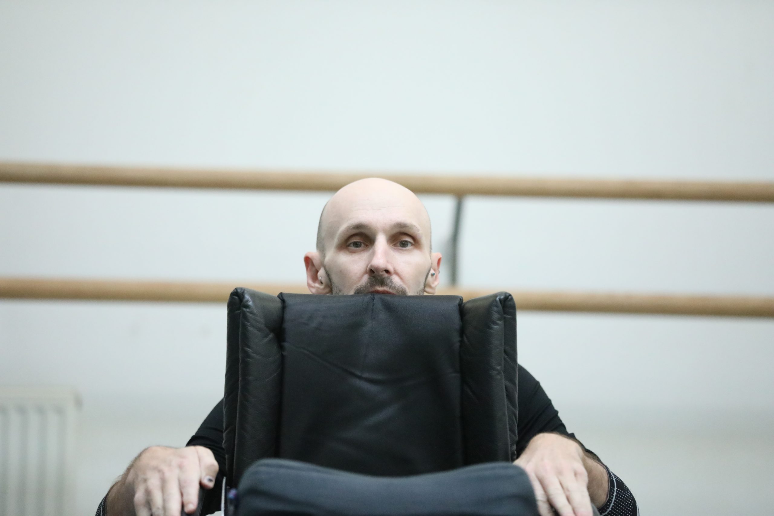 In a dance studio, Marc peers over the top of the back of his black wheelchair, hands extended onto the arms of the chair.