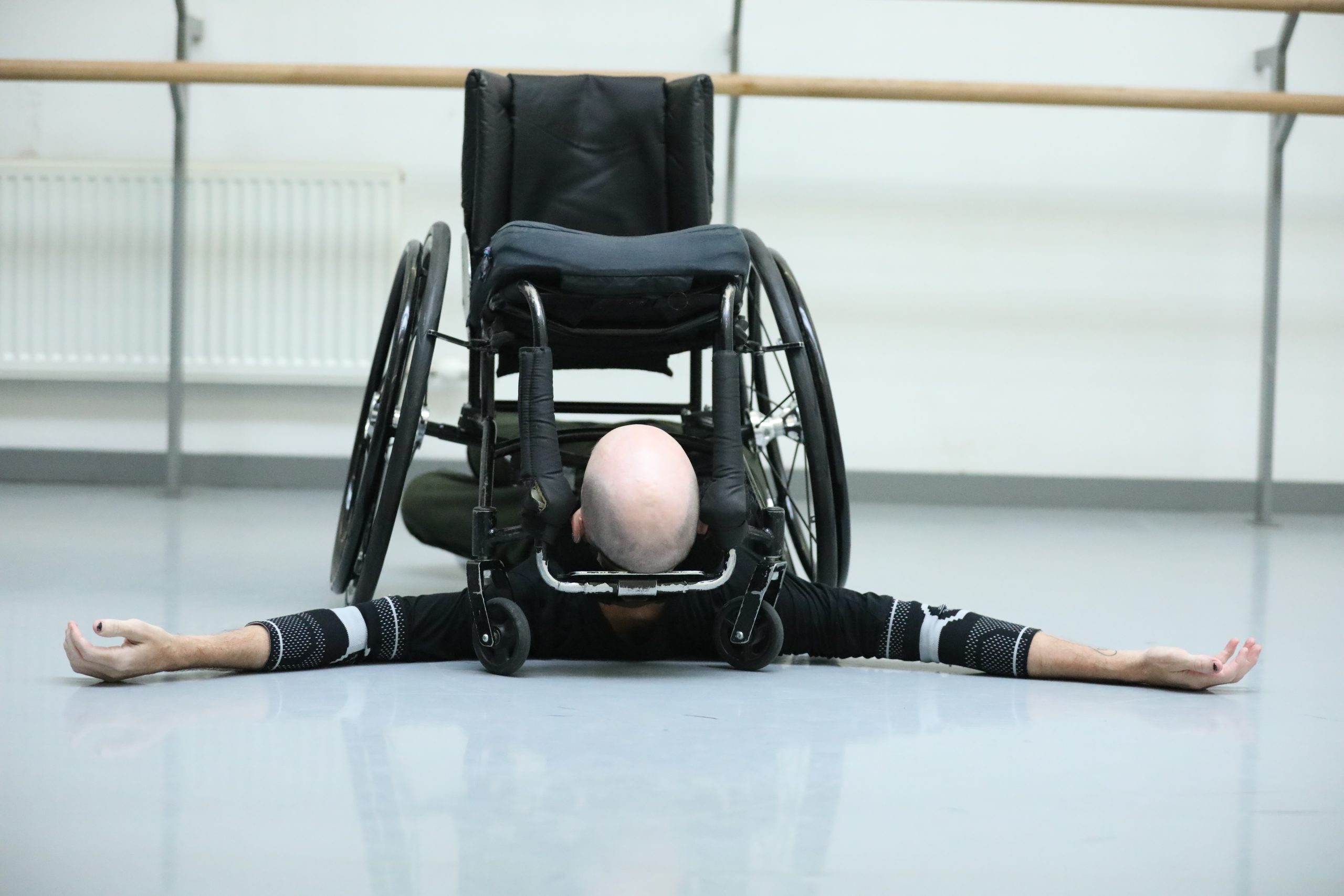 In a dance studio, Marc is laid flat on the floor, his arms stretched out above his head. His wheelchair sits on top of him, trapping his head between the footrest and his arms between the wheels.
