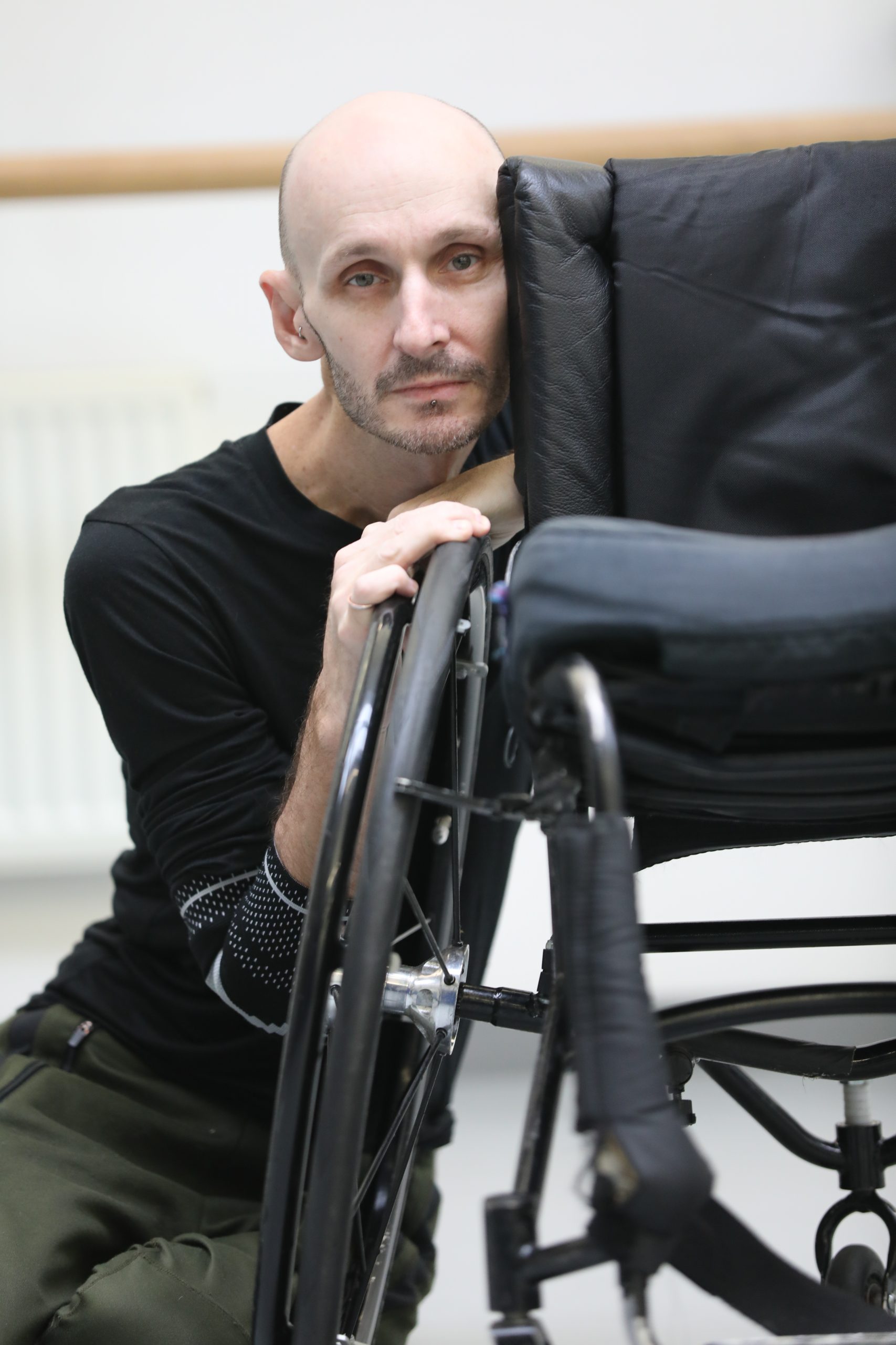 From seated on the floor Marc leans his body against his wheelchair, holding onto the wheel.