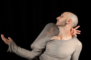 Marc (dancer) is in a grey long sleeved top, his arm out stretched. Caroline (dancer) has grey gauze stretched over her face. she reaches her arms round Marc's neck and chest. They are cropped at the torso, on a black background.