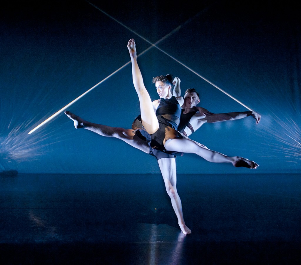 Rebecca Evans and James Cousins, dancers on a dark stage. Legs and arms reaching outwards, white likes blurring above their heads.