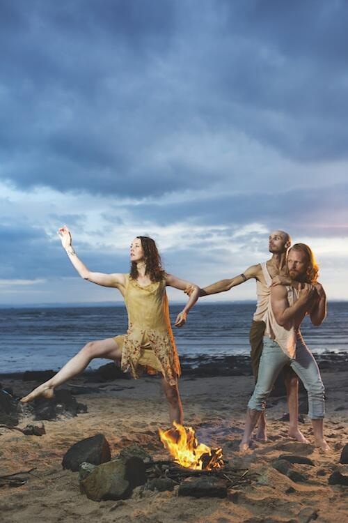Three castaways are on a beach by a fire. Two men hold the arm of a woman as she reaches away from them.