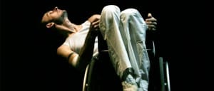 Marc leans back away from us at an angle in his wheelchair. He wears all white clothing against a black background.