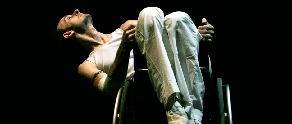 Marc leans back away from us at an angle in his wheelchair. He wears all white clothing against a black background.