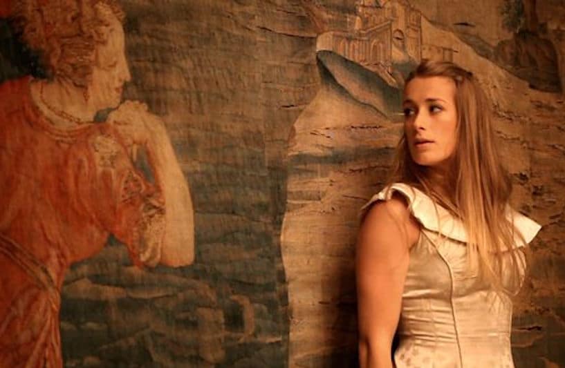 A dancer with long blonde hair, in a fitted cream dress with a bodice, leans against a faded tapestry.