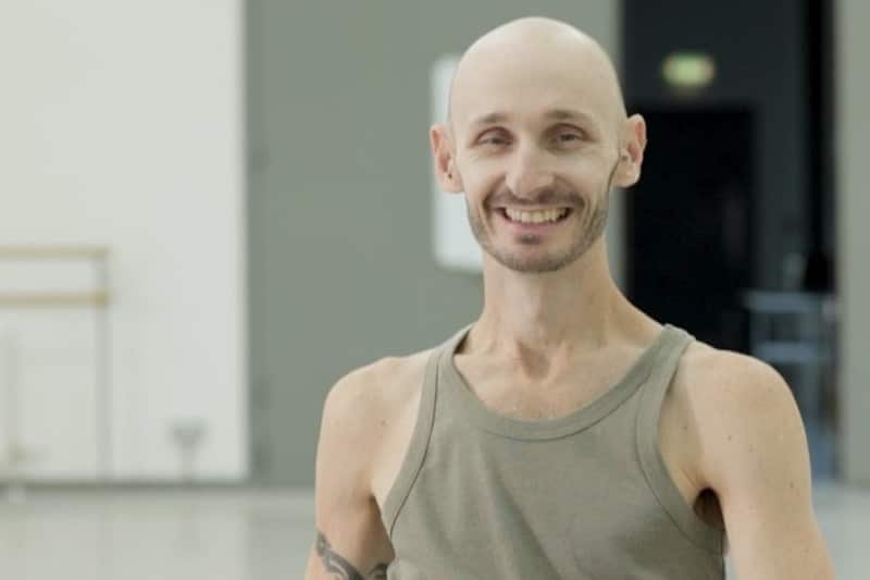 A portrait of Marc Brew smiling in a dance studio. Wearing a grey vest.