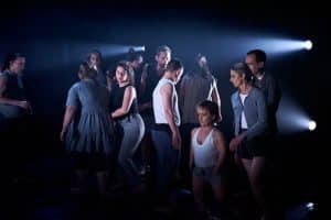 An ensemble of dancers are gathered together on a dark stage, spotlit by rays of light