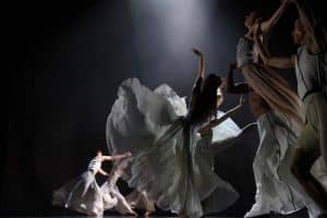 An ensemble of dancers move across a dark background. Large white, transparent skirts fan out around them