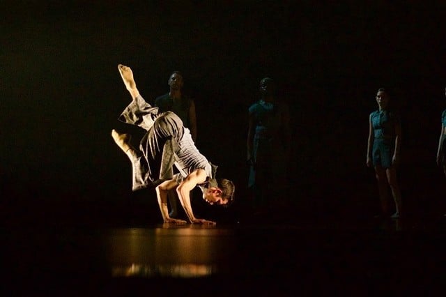 A dance spotlit on a dark stage balances on their hands, their legs raised up above them.