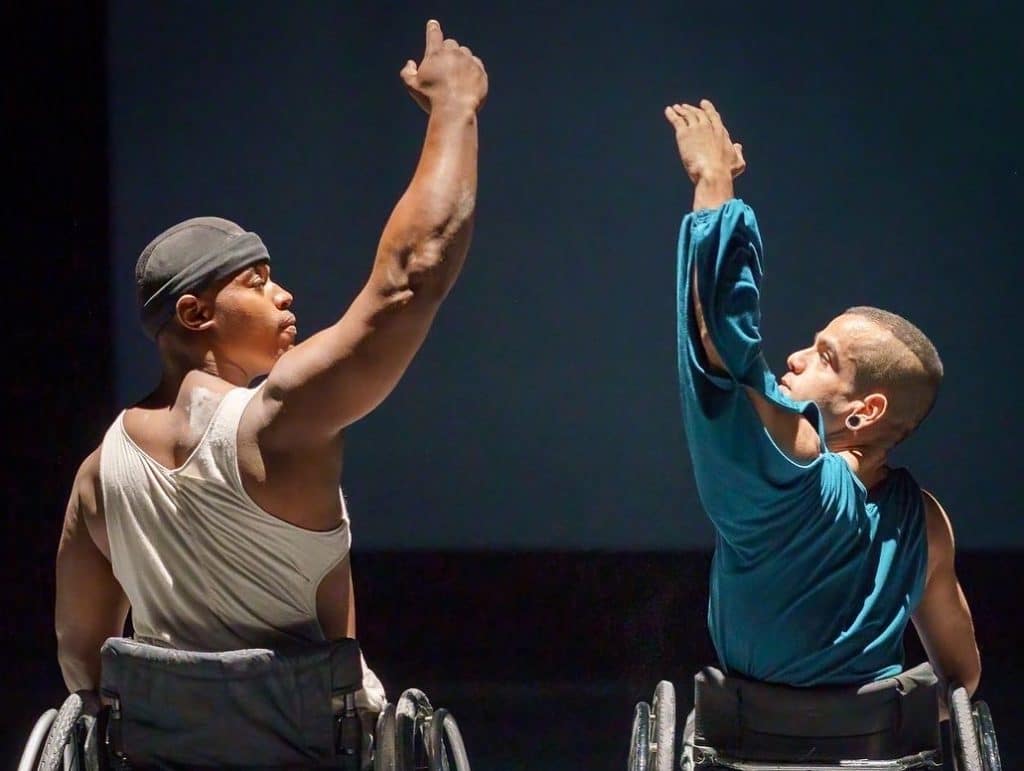 Two dancers in their wheelchairs face each other side on. Their arms are raised.