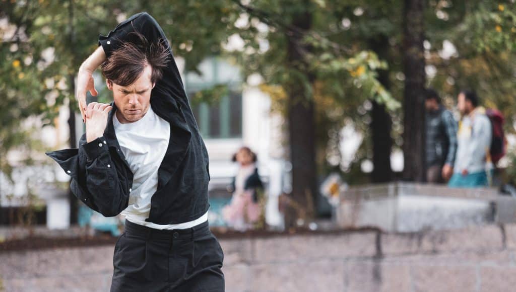 A dancer dressed in black jeans and denim jacket and white tishirt dances on a city street infront of trees. One arm reaches over his head to grasp the other hand.