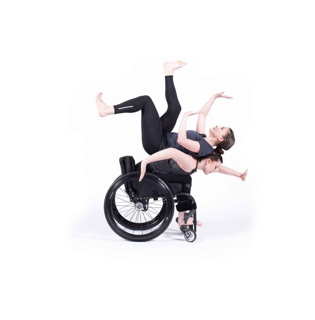 A dancer balances on the back of a fellow dancer in a wheel chair. They counter balance each other by reaching out arms and legs. They are dressed in black against a bright white background.