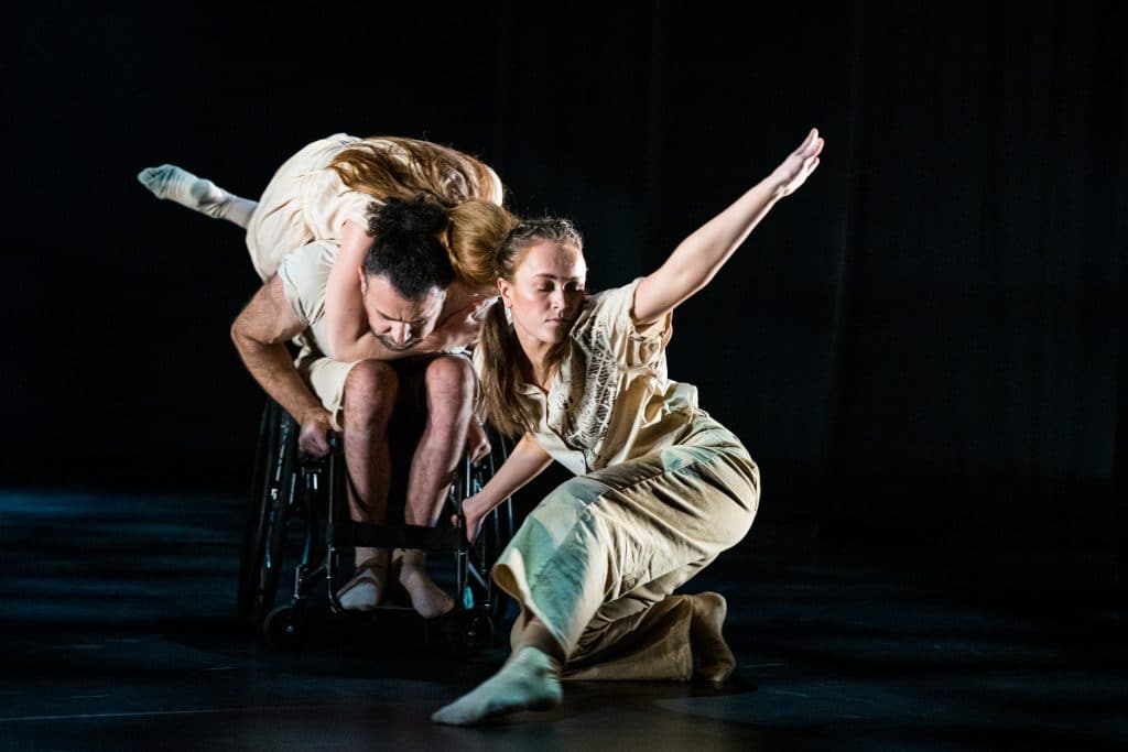 A dancers grasps the neck of another dancer in a wheelchair, she lies across his back. Another dancer crouches infront of them an arm outstretched to the sky. They are surrounded by black.
