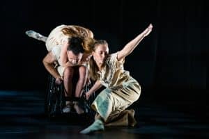 A dancers grasps the neck of another dancer in a wheelchair, she lies across his back. Another dancer crouches infront of them an arm outstretched to the sky. They are surrounded by black.