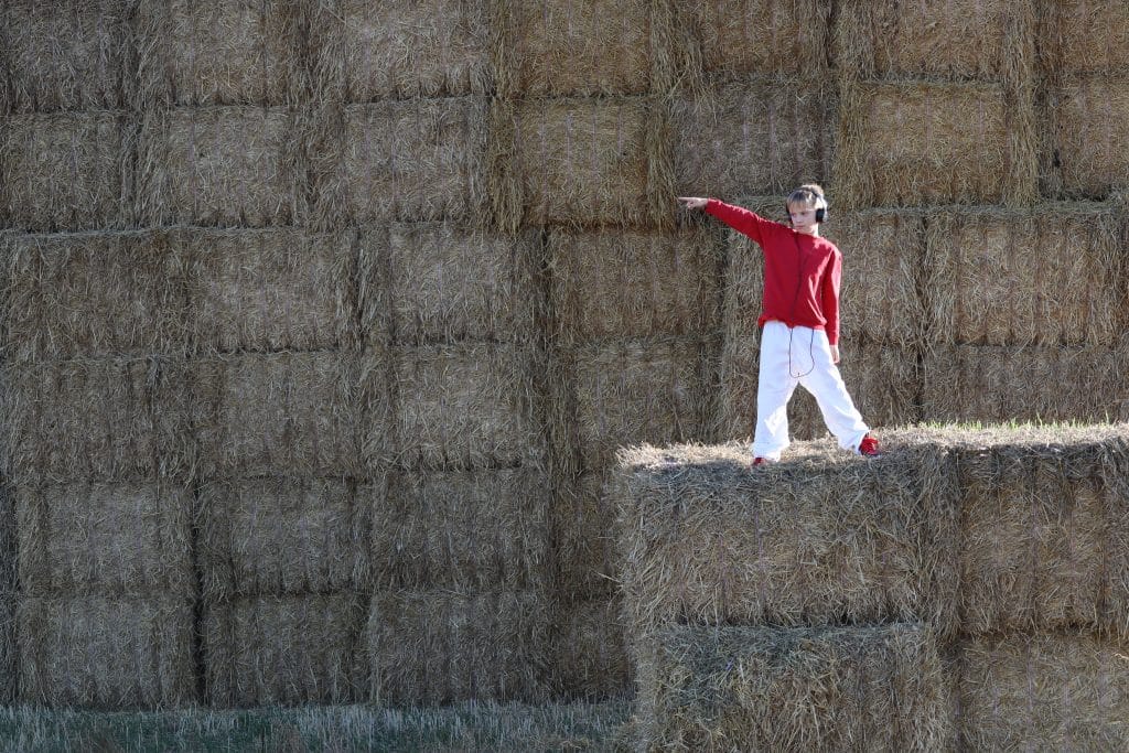 A blonde hair boy wearing a red jumper, white trousers, and black head phones poses mid dance on haybales. He points and looks off towards his right. His backdrop is haybales filling the image.