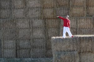 A blonde hair boy wearing a red jumper, white trousers, and black head phones poses mid dance on haybales. He points and looks off towards his right. His backdrop is haybales filling the image.