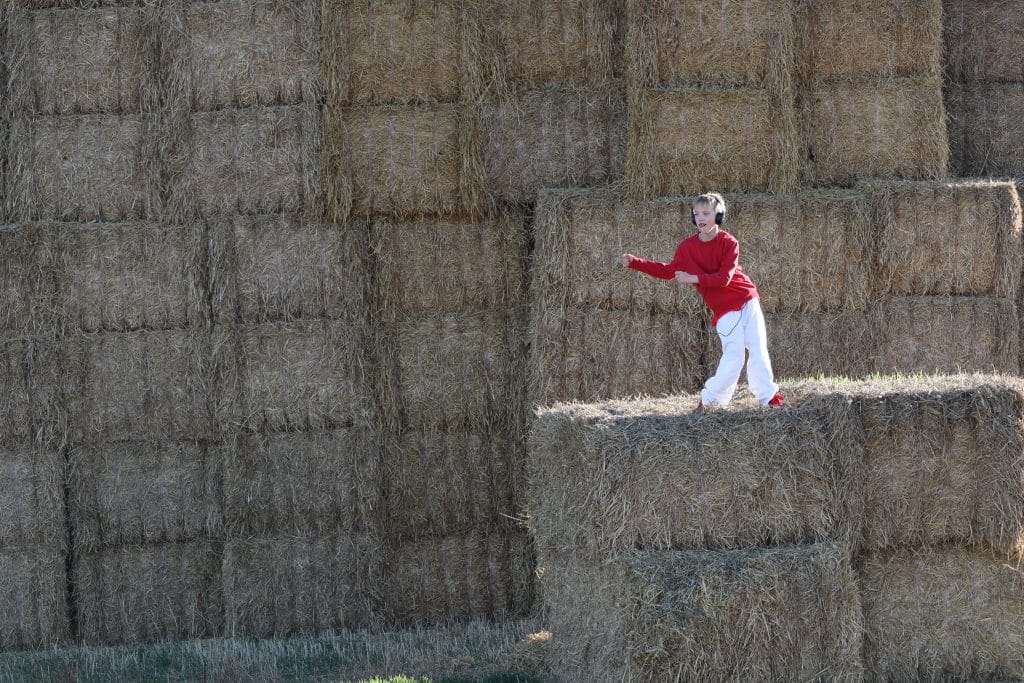 A young blonde haired boy wearing white trousers, a red jumper and large black headphones dances on a hay stack