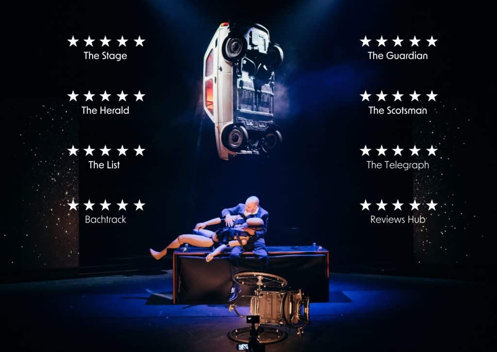 Marc sits on a desk in the middle of a stage holding a crash dummy, a wheelchair on its side lies at the front of the stage and a car is suspended over him. Multiple 5 star reviews listed down the sides of the image from The Stage, The Guardian, The Herald, The Scotsman, The List, The Telegraph, Bachtrack and Reviews Hub.