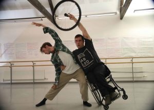 A shot of Ross and Piotr dancing in a studio. Piotr leans into Ross and lifts his wheel of his wheelchair over his head. Ross also leans into Piotr with his arm stretched over his head.
