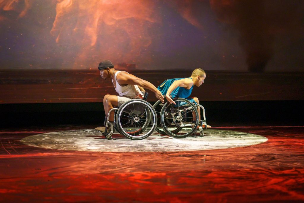 Two dancers in wheelchairs are centre stage. They have their backs to each other, leaning forward with their arms pushed behind them. Their hands are facing each others. There is a circular spot light on them with red and orange lighting and backdrop.