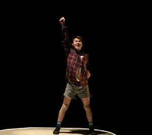 A dancer is on a dark stage, with a spotlight on him. Hw is wearing a checkered shirt and denim shorts. He is standing on a wooden ramp. He has one hand on his hip and another is pushing up to the sky.