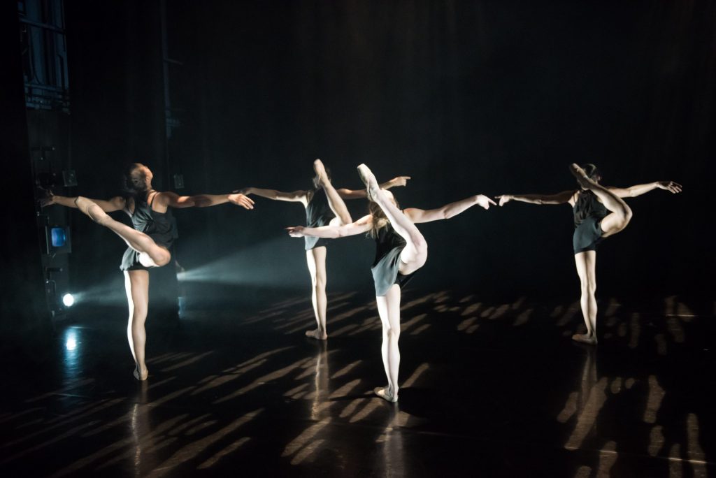 4 dancers are on a darkened stage. They are all facing the back with their arms straight out to the sides and one leg lifted up in the air behind them, toes pointed to the ceiling.