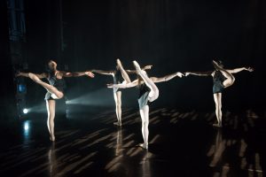 4 dancers are on a darkened stage. They are all facing the back with their arms straight out to the sides and one leg lifted up in the air behind them, toes pointed to the ceiling.