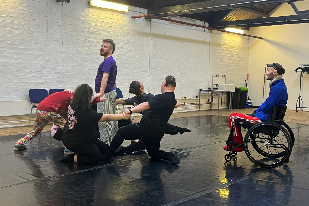 In a dance studio Independance ensemble are in mid movement. Dancers hold hands kneeling in a circle with one dancer in the middle. Marc is in his wheelchair watching them.