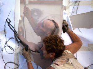 Rachel sketches on a large white cloth outdoors. The sun shines through the fabric and we can see an outline of Marc.