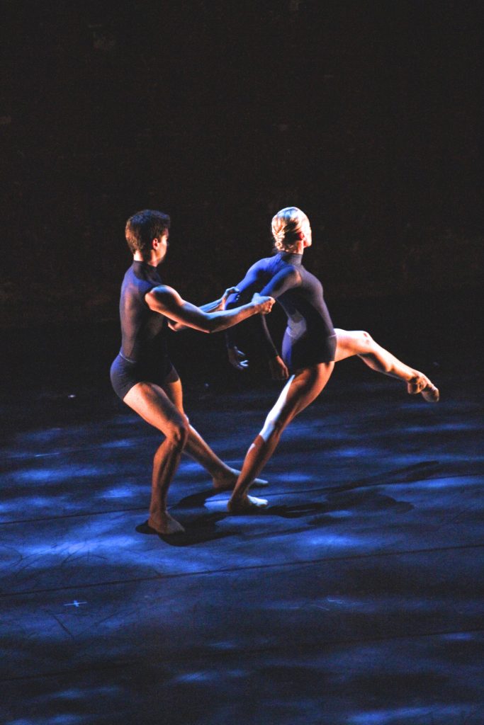 Two dancer are on stage in dark leotards, tops and shorts. One dancer has their back tot he other. Their arms stretched behind them and the other dancer holds on. The dancer facing away has one leg stretched and pointed out in front of them.