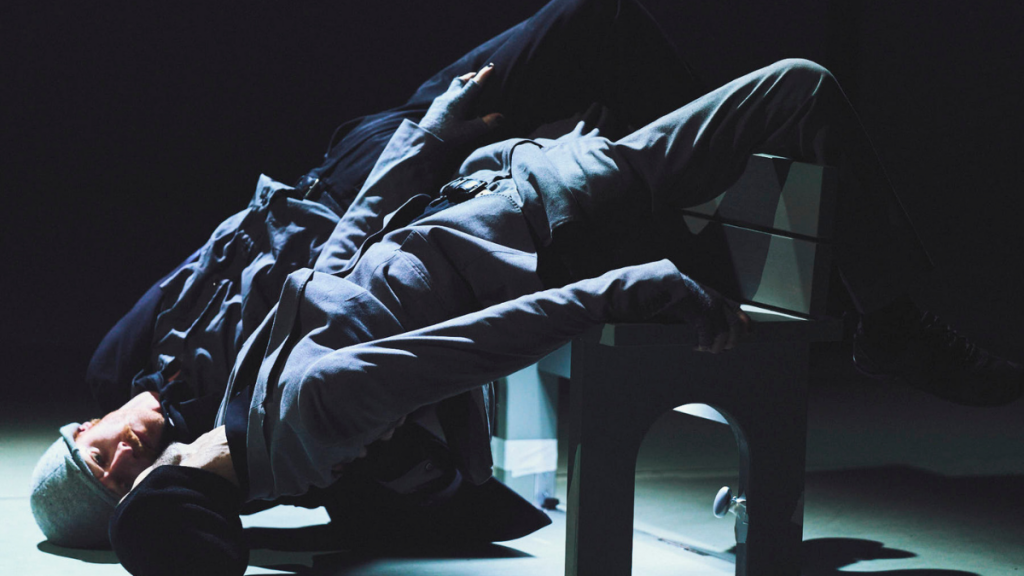 Marc And Nelson are backwards and leaning over a bench. Their heads are on the floor with their legs over the back of the bench, looking at each other. They are both wearing suits and a grey hat.