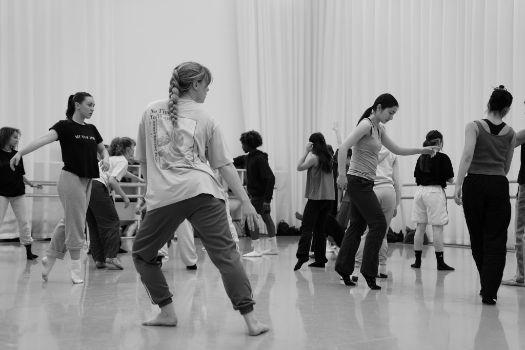 Hayley is in a large dance studio with curtains all along the walls and ballet barres. She is in mid movement with an ensemble of dancers around her also in various movement positions. The image is in black and white.