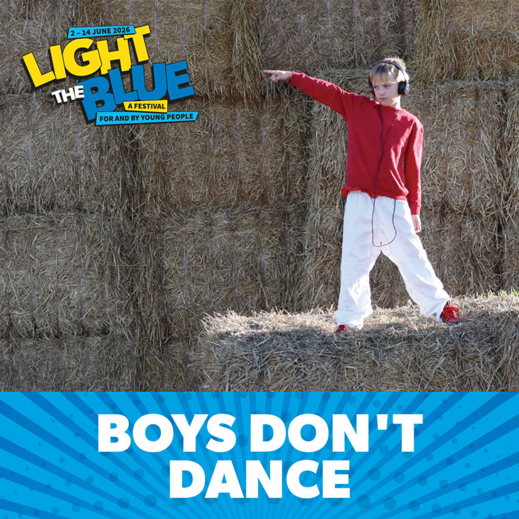 A blonde hair boy wearing a red jumper, white trousers, and black head phones poses mid dance on haybales. He points and looks off towards his right. His backdrop is haybales filling the image. This has a banner at the bottom of the image with blue stripes and says "Boys Don't Dance". The Light the Blue Festival logo is in the left top corner.