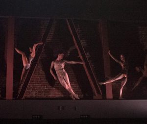 3 dancers lean onto metal beams, with a large red bricked wall behind them.