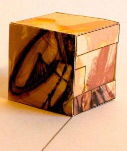 A cube with drawings and sketches in white, beige and orange tones, sits on a white piece of paper. On the most visible face of the cube you can see the sketch of wheelchair wheels.