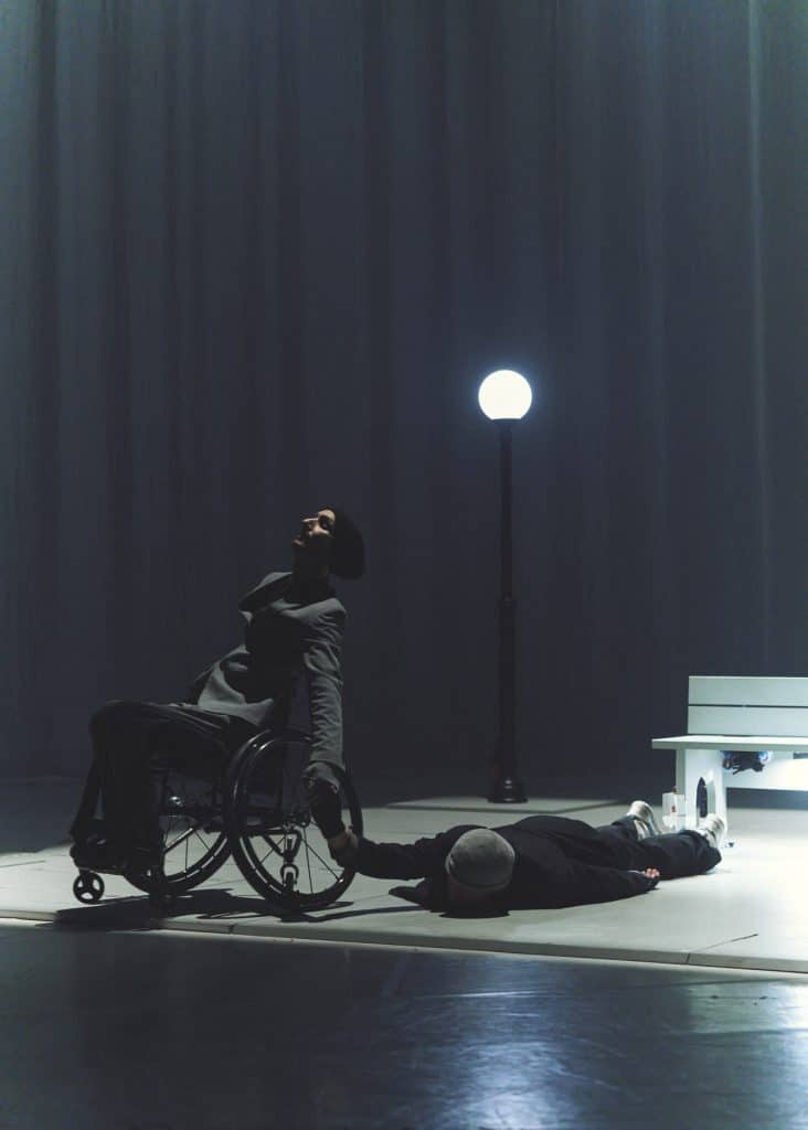 Marc and Nelson hold hands, Marc is in his wheelchair, while Nelson is lying flat on the floor. A bench and a street lamp are in the background. They both wear suits and grey hats.