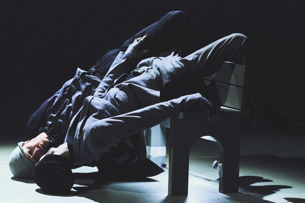 Marc And Nelson are backwards and leaning over a bench. Their heads are on the floor with their legs over the back of the bench, looking at each other. They are both wearing suits and a grey hat.