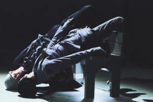 Marc And Nelson are backwards and leaning over a bench. Their heads are on the floor with their legs over the back of the bench, looking at each other. They are both wearing suits and a grey hat.