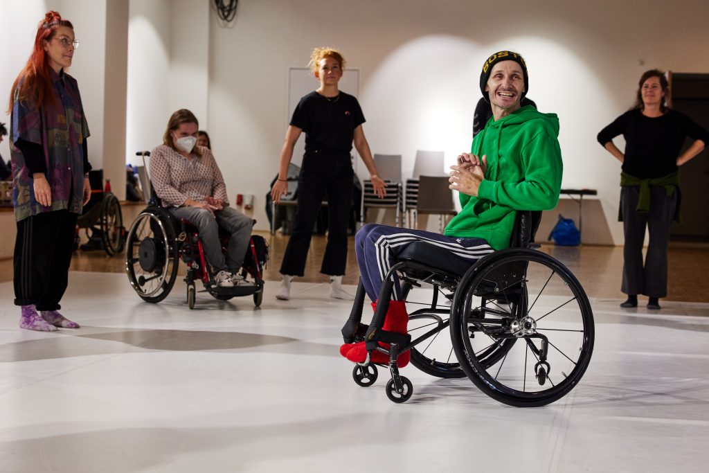 Marc Brew is in his wheelchair in a dance studio with other dancers in a circle. He has a green jumper on and smiling.