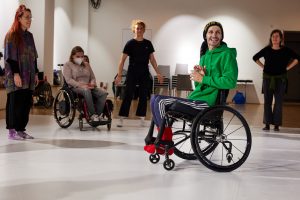 Marc Brew is in his wheelchair in a dance studio with other dancers in a circle. He has a green jumper on and smiling.