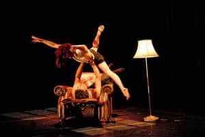 A dark stage with a warm spotlight on two dancers. There is a large armchair and floor lamp on stage. One dancer is lying on the arm chair, the other dancer is in mid air, the dancer holding them above them, with their arms and arms spread out straight.