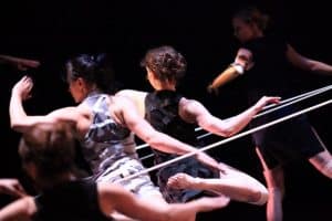 A close up shot of an ensemble of dancers on stage mid movement. There are poles attached to the dancers sides.