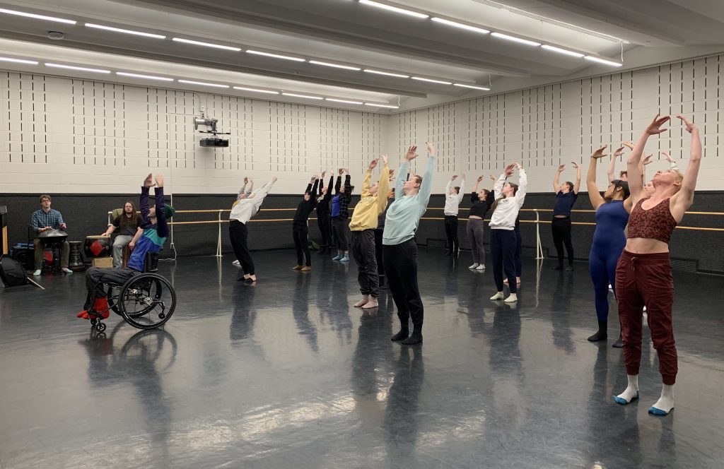 In a dance studio with dark floors, white walls and a ballet barre around the walls. Marc is in his wheelchair at the front of the studio demonstrating. He has his arms above his head, leaning back with his gaze to the left. The group of dancers behind him are in the same position.