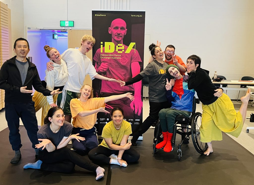Marc and a group of dancers are posing in front of a large pink and yellow poster. That has an image of Marc on with text saying"IDEA Independent Dance Exchange Australia". The group are all smiling in different poses.
